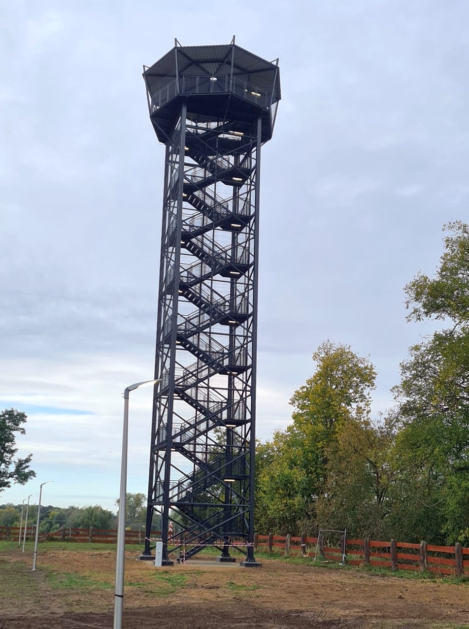 Wysoka, metalowa wieża widokowa z zewnętrznymi schodami spiralnymi, otoczona drzewami i oświetlona przez nowoczesne latarnie.