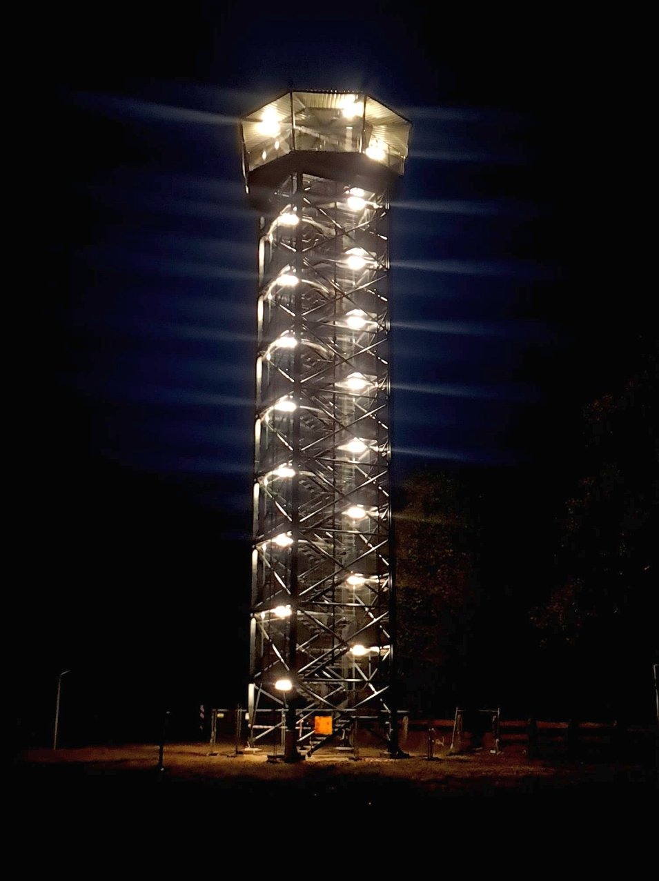 Oświetlona, wysoka wieża widokowa ze stalową konstrukcją i schodami, sfotografowana nocą.