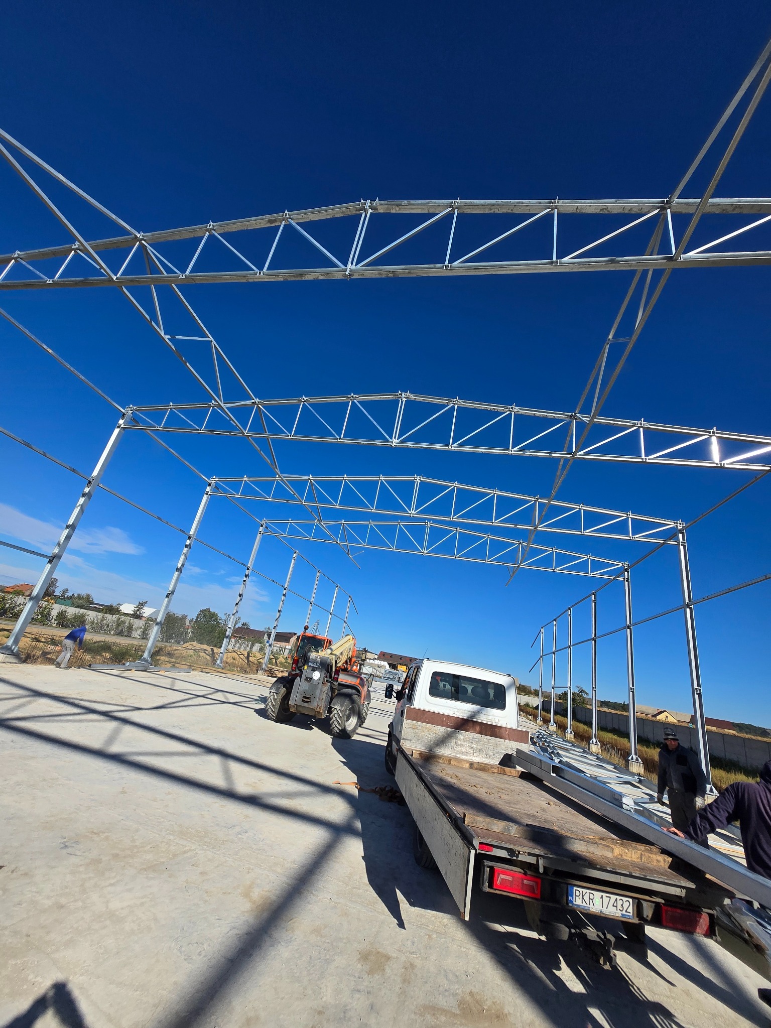 Stalowa konstrukcja hali w trakcie montażu, widoczny podnośnik teleskopowy i samochód dostawczy z elementami konstrukcyjnymi.