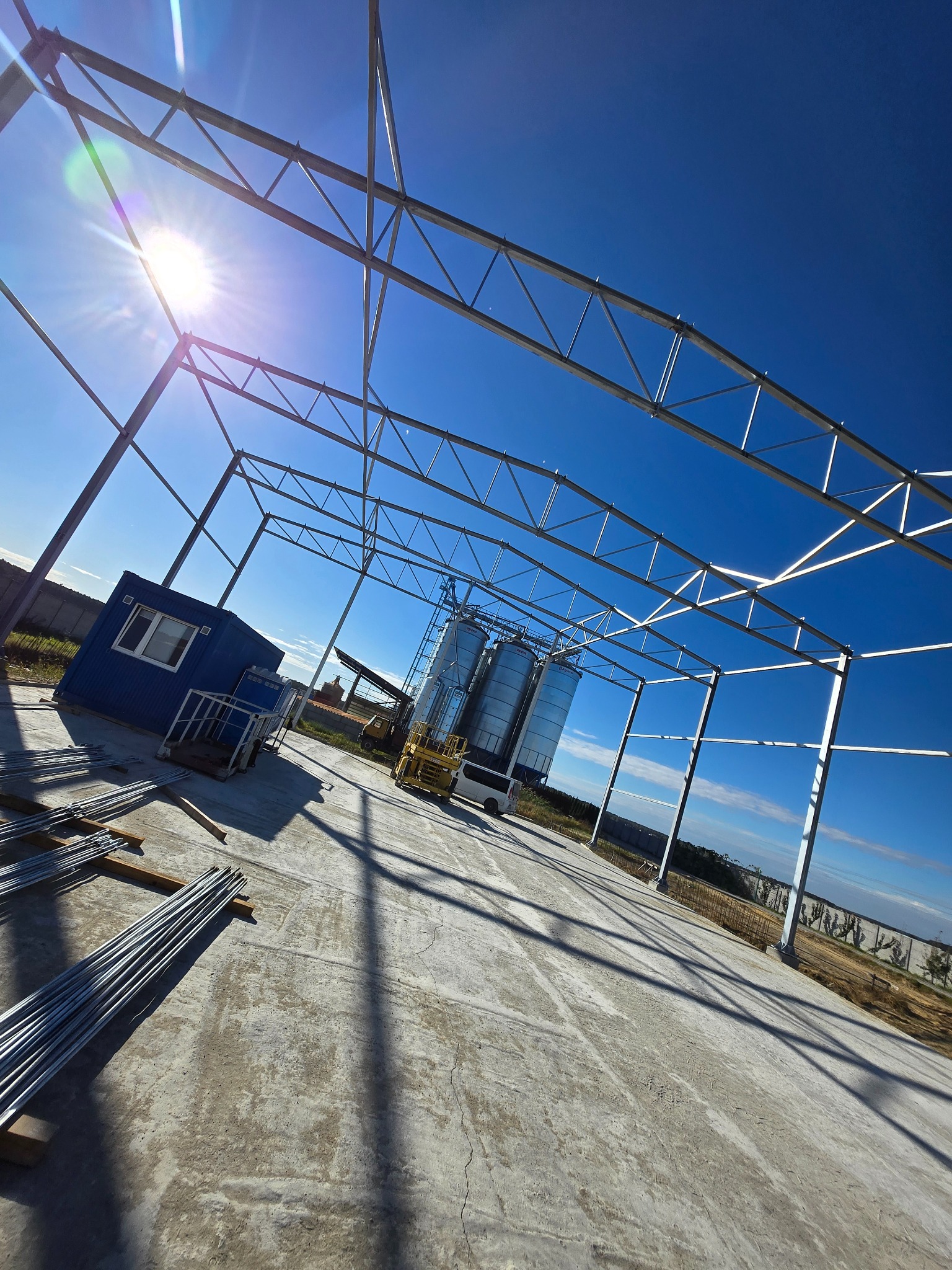 Konstrukcja stalowa hali w trakcie budowy, widoczne elementy szkieletu, niebieski kontener biurowy i silosy w tle, jasne słońce.