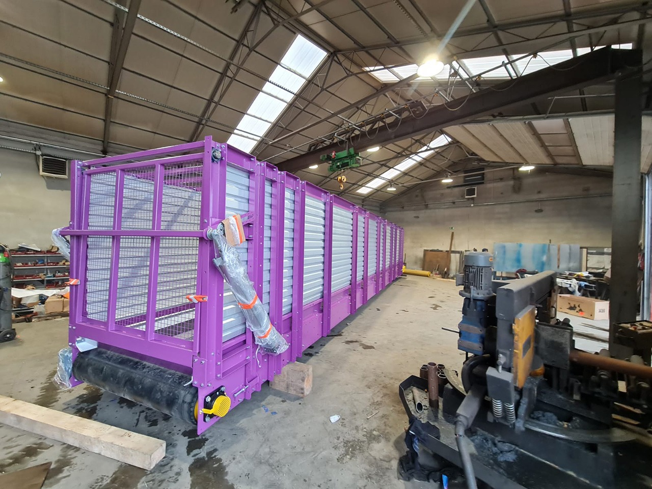 Długa, fioletowa konstrukcja stalowa z kratownicą i elementami transportowymi na hali produkcyjnej, widoczne narzędzia i suwnica.