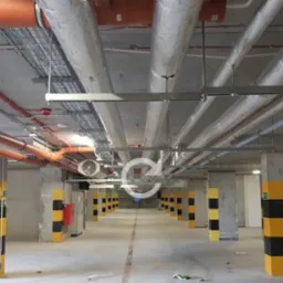 Instalacje wentylacyjne i rury poprowadzone pod sufitem parkingu podziemnego, widoczne kolumny z oznaczeniami ostrzegawczymi.