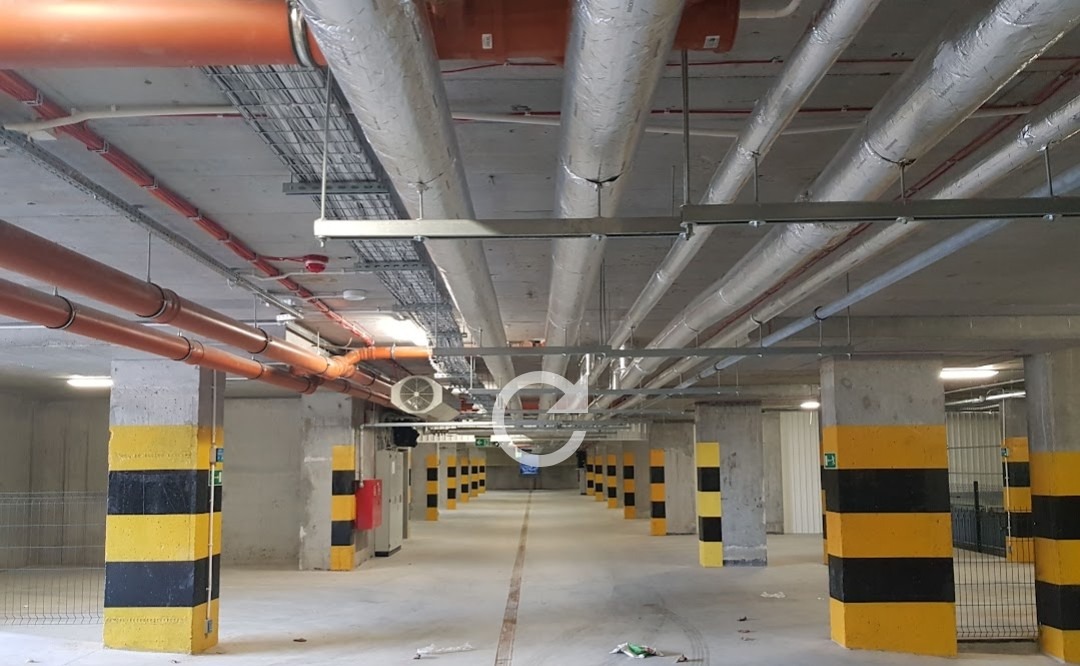 Instalacje wentylacyjne i rury poprowadzone pod sufitem parkingu podziemnego, widoczne kolumny z oznaczeniami ostrzegawczymi.