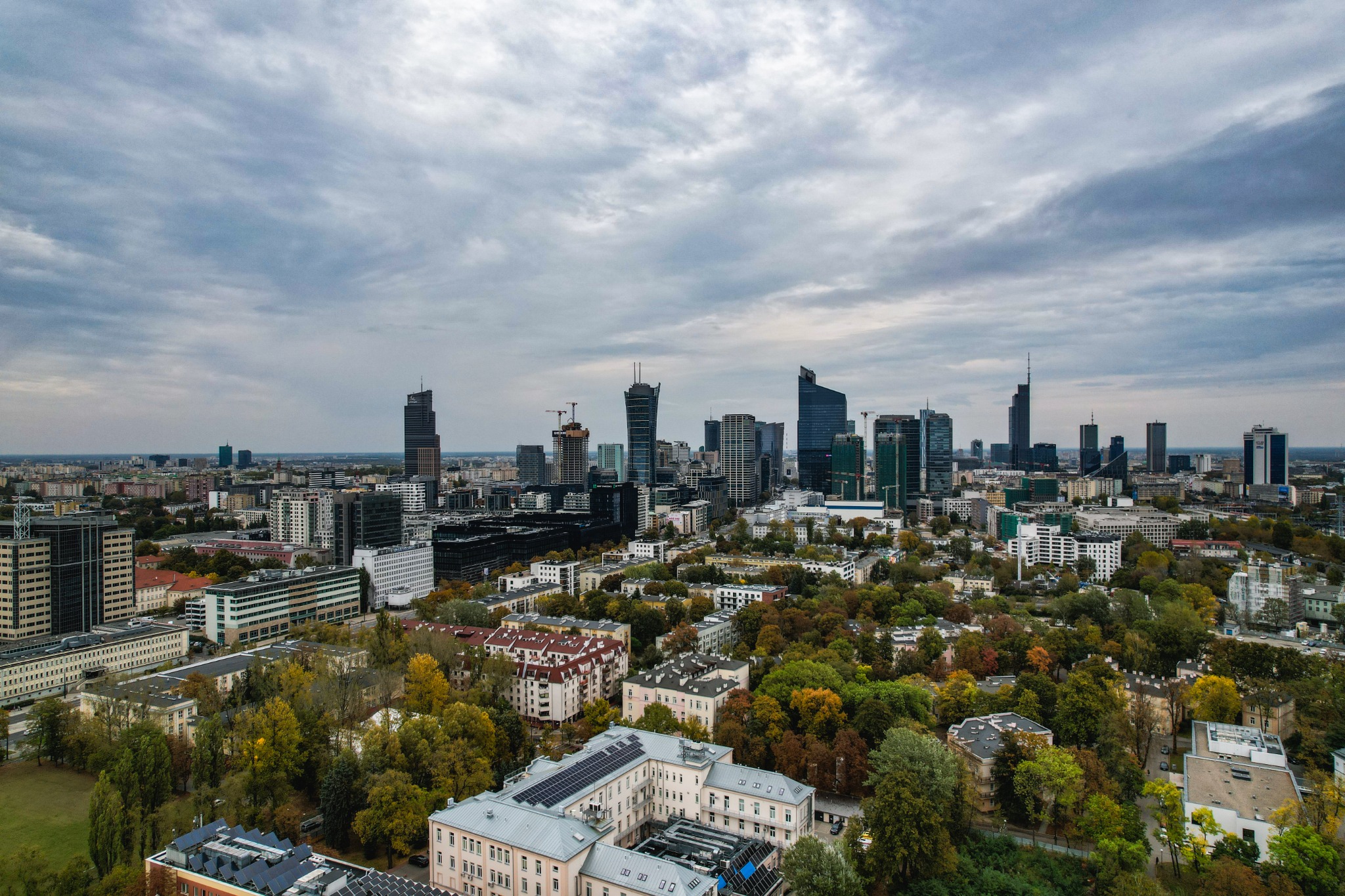 Panorama Warszawy z widokiem na wieżowce w pochmurny dzień, zieleń drzew w centrum kadru.