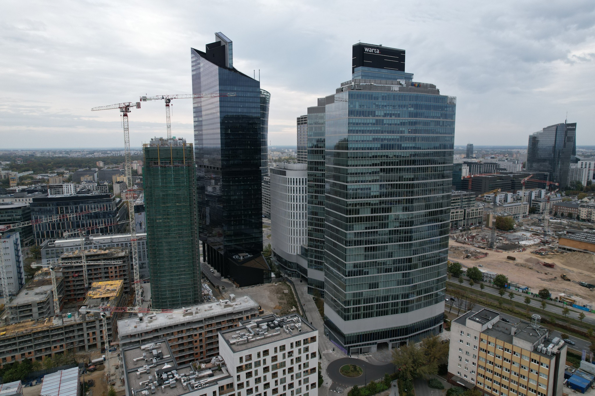 Panorama Warszawy z wysokości, ukazująca nowoczesne wieżowce, w tym budynek z logo Warta na szczycie, oraz budynki w budowie z żurawiami.
