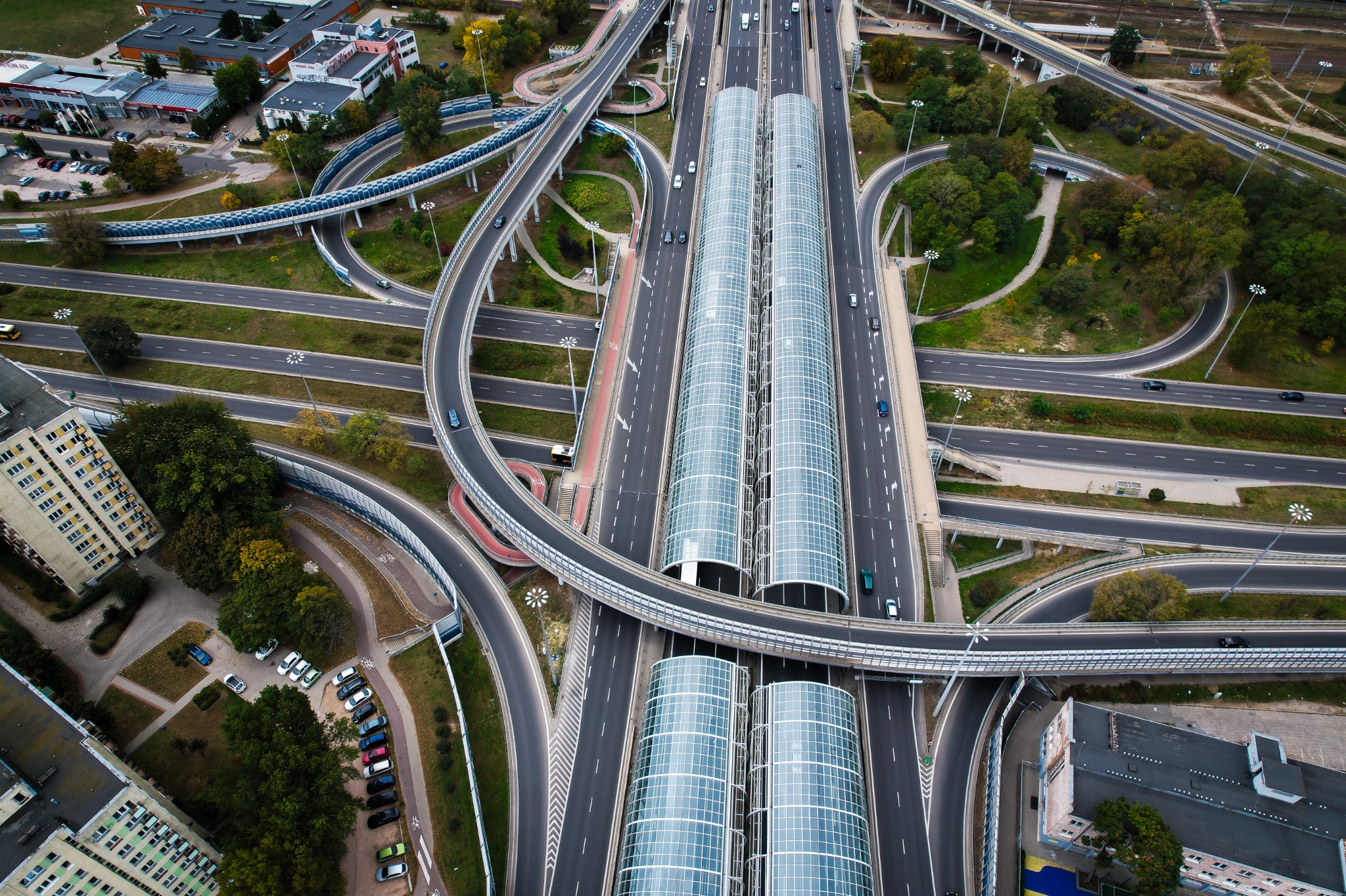 Widok z lotu ptaka na skrzyżowanie autostrad z charakterystycznymi zadaszonymi przystankami autobusowymi, otoczonymi zielenią i budynkami.