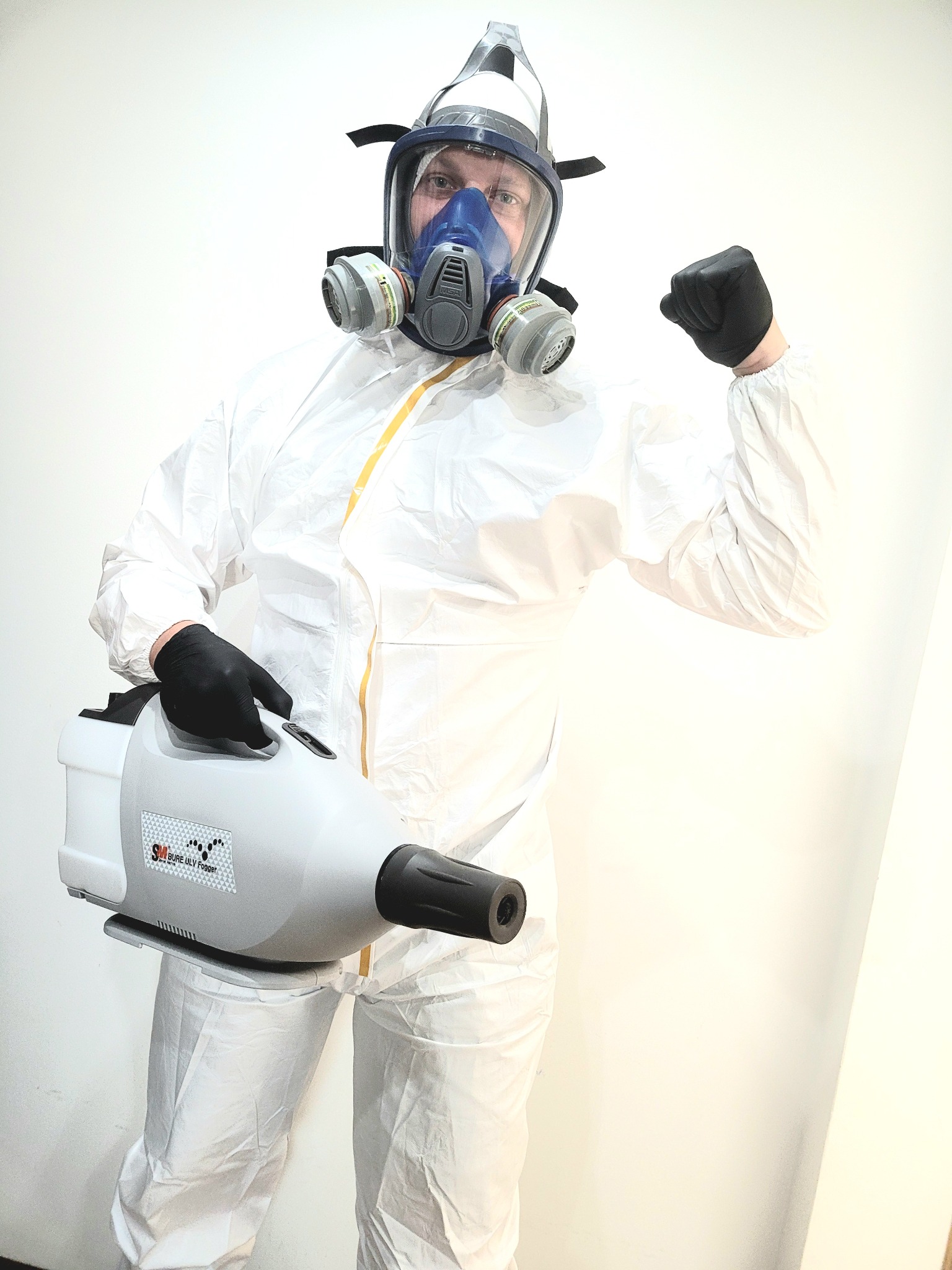 Osoba w białym kombinezonie ochronnym i masce gazowej, trzymająca zamgławiacz Sure ULV Fogger i prezentująca zaciśniętą pięść na tle jasnej ściany.