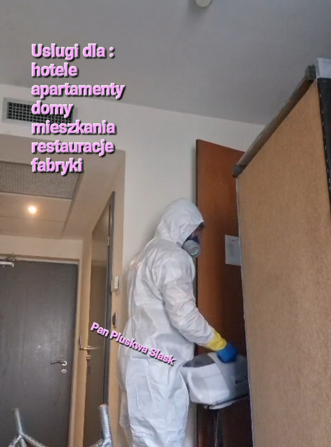 Pracownik w kombinezonie ochronnym i masce, z niebieskimi rękawicami, przeprowadza zabieg z użyciem specjalistycznego urządzenia w pokoju hotelowym, obok widoczna szafa i drzwi.