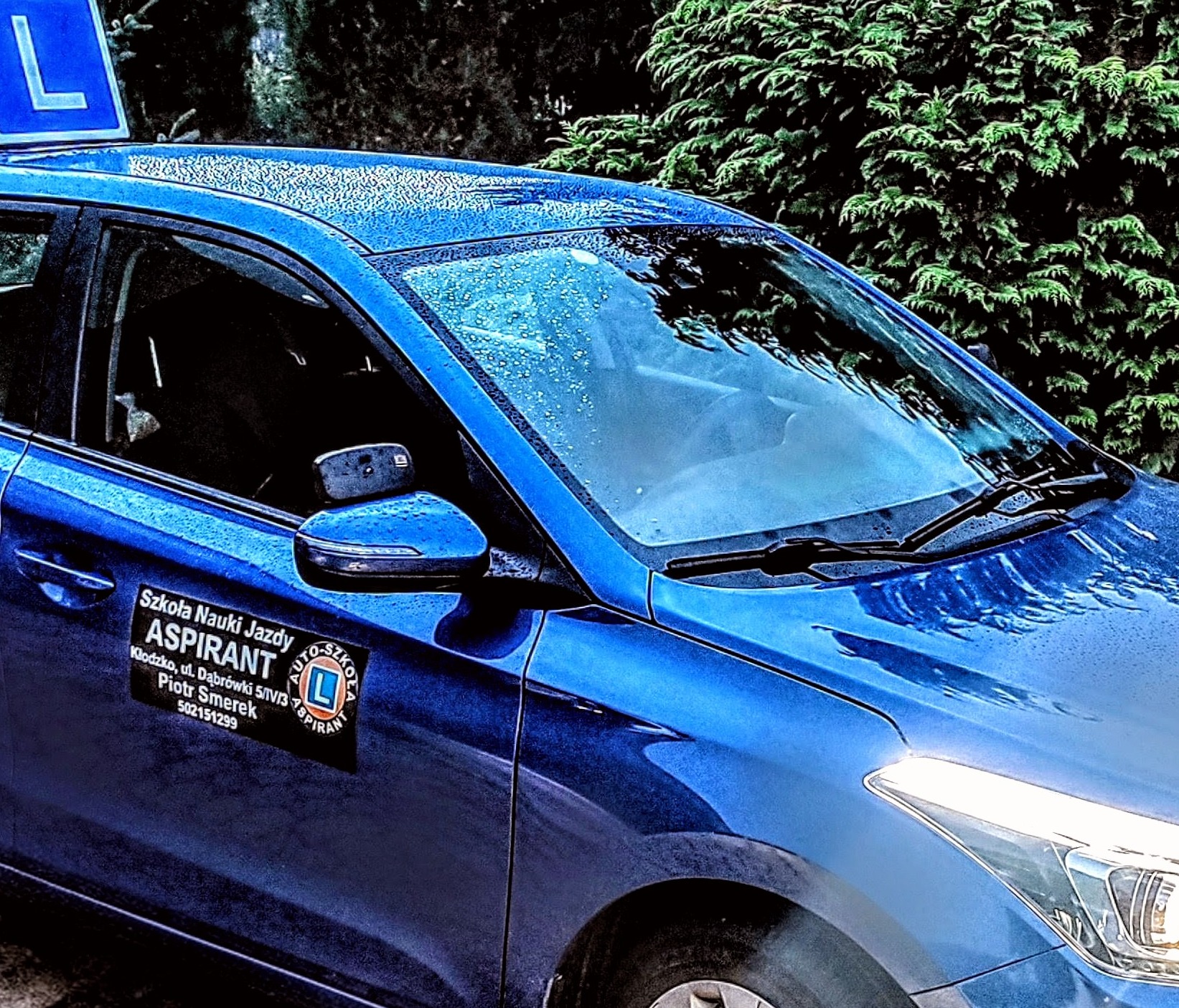 Niebieski samochód oznaczony literą 'L' na dachu, z naklejką 'Szkoła Nauki Jazdy Aspirant' na drzwiach, zaparkowany na tle zieleni.
