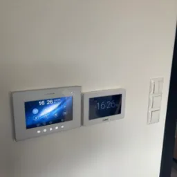 Dwa panele sterowania domofonem Vidos i Satei zamontowane na białej ścianie, obok włącznik światła i fragment czarnej futryny drzwiowej. Ekrany pokazują godzinę 16:26.