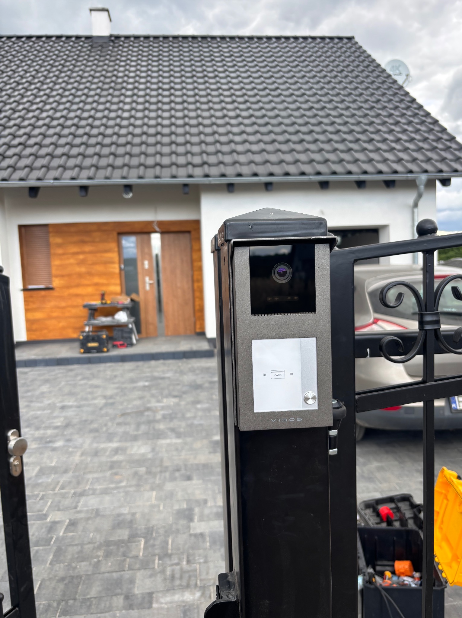 Nowoczesny wideodomofon Vidos zamontowany na czarnym słupku bramy wjazdowej. W tle widoczny dom z grafitowym dachem i drewnianą elewacją. Skrzynka narzędziowa obok.