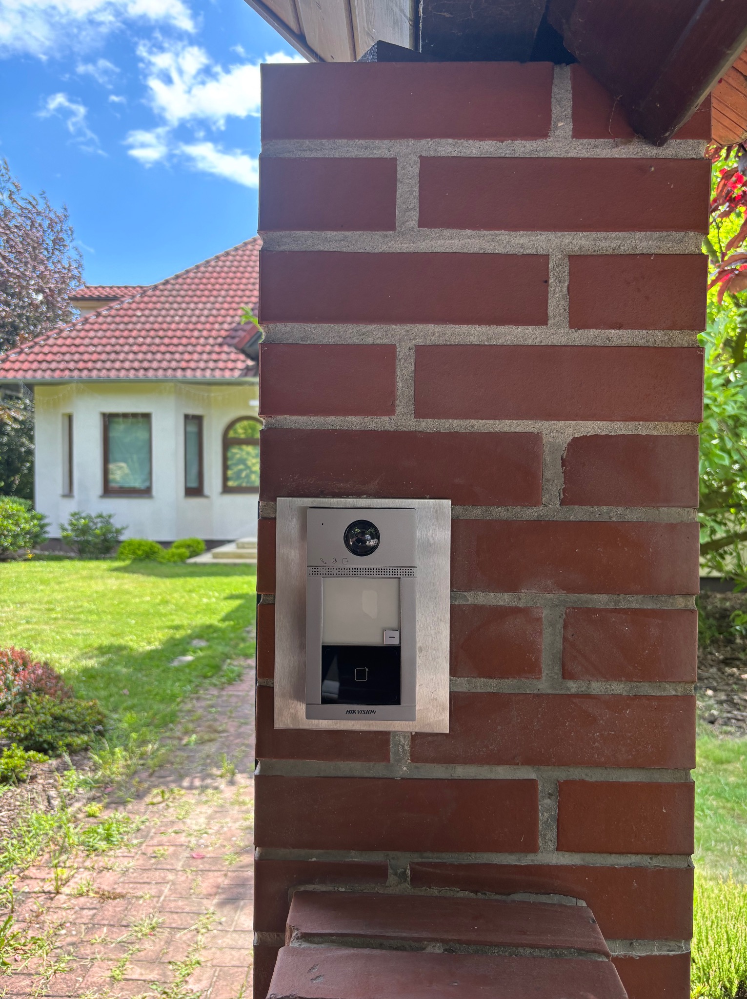 Nowoczesny wideodomofon Hikvision zamontowany na ceglanym słupku przy wejściu do domu z zielonym ogrodem w tle, widok z dołu.