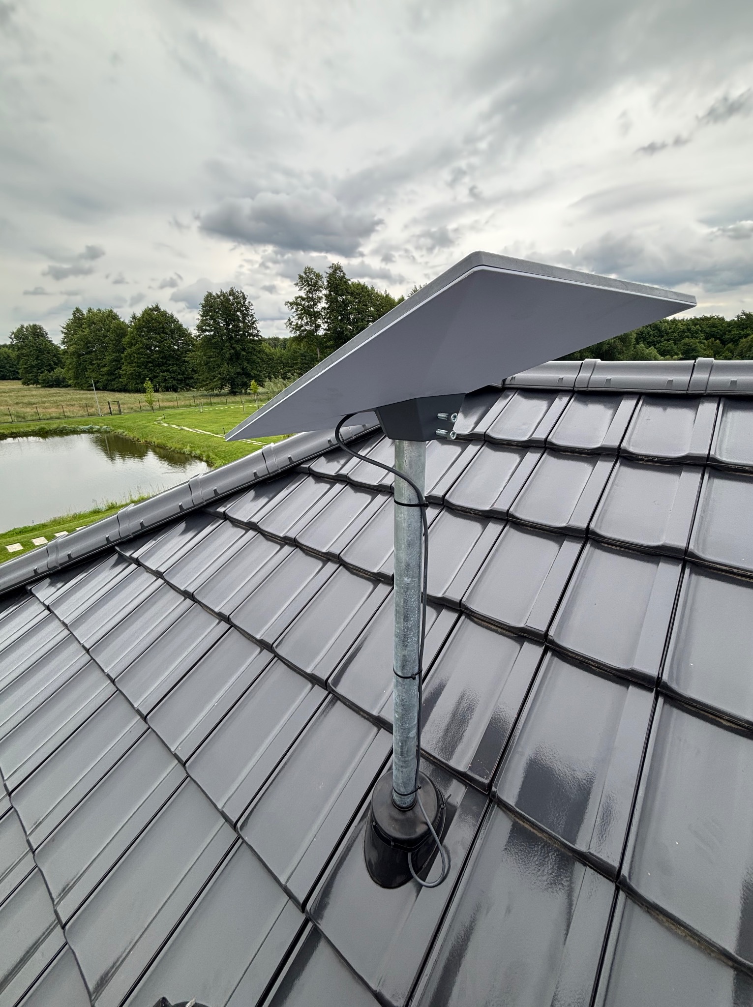Antena satelitarna Starlink zamontowana na metalowym maszcie na dachu z czarnej dachówki. W tle widok na zielone drzewa i małe jezioro pod pochmurnym niebem.