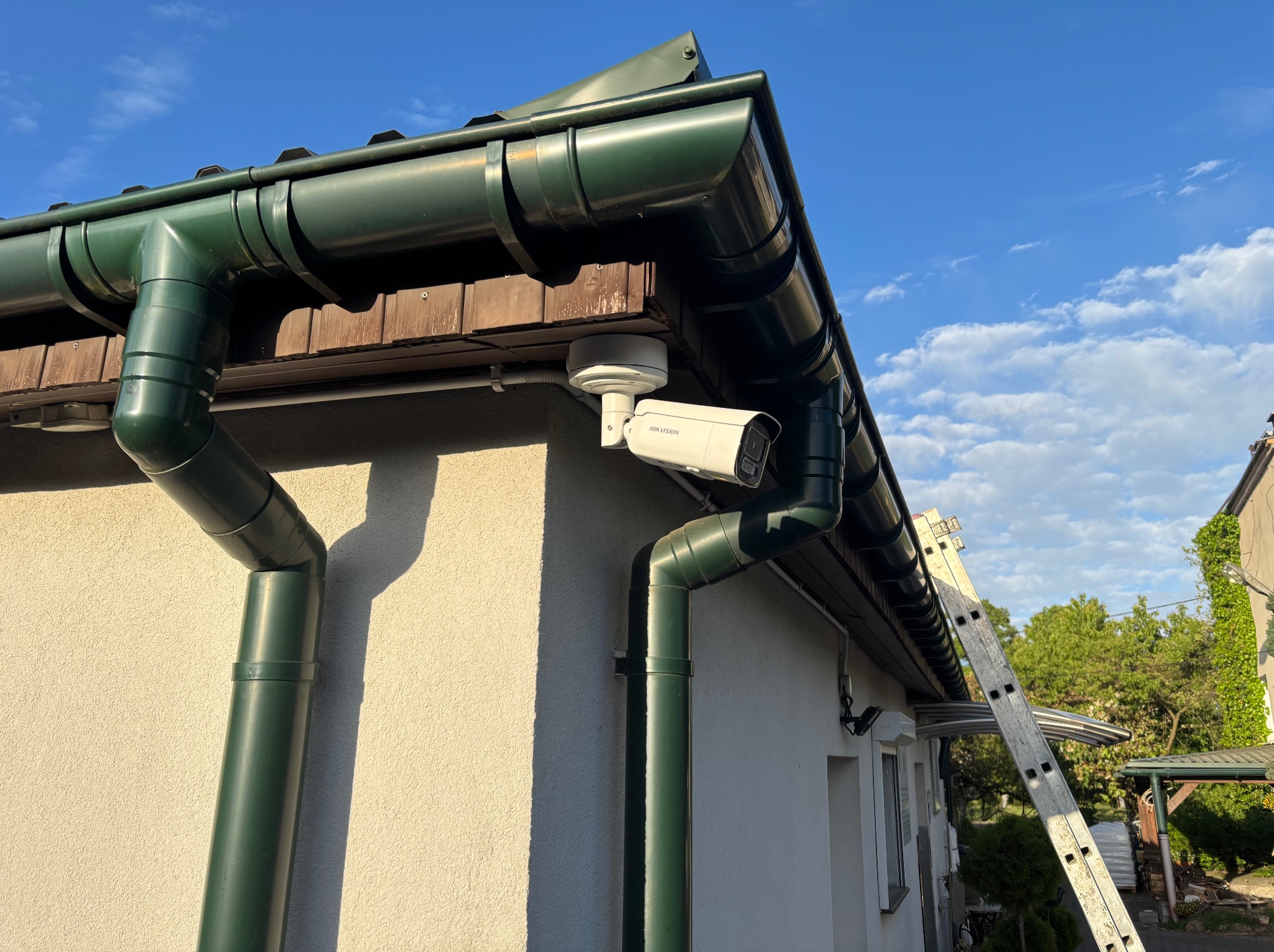 Kamera Hikvision zamontowana na elewacji budynku pod zieloną rynną. Widoczny fragment drabiny oparty o budynek. Błękitne niebo w tle.