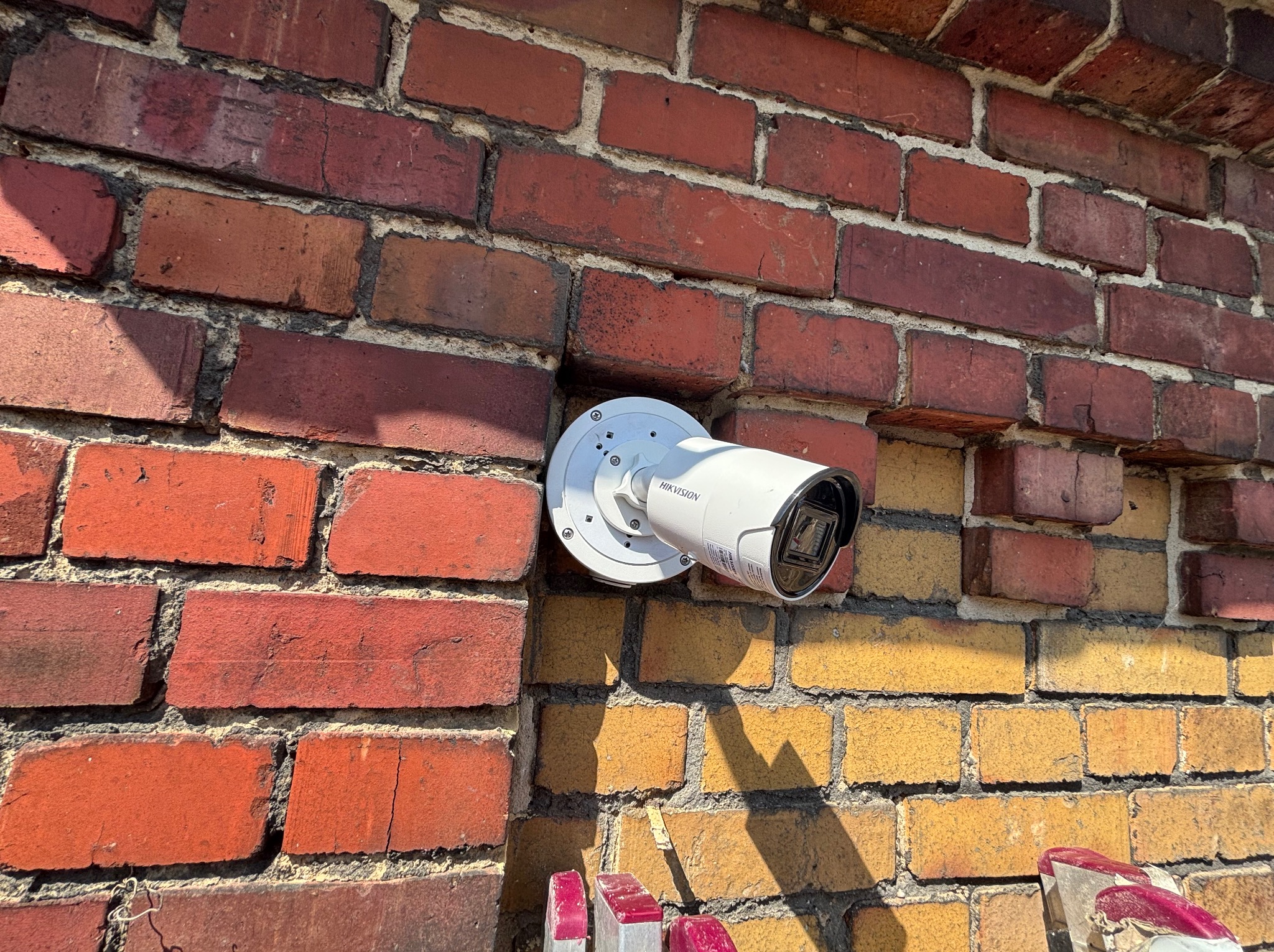 Kamera monitoringu Hikvision zamontowana na ceglanej ścianie zewnętrznej budynku, widoczna z dołu pod kątem, w jasnym oświetleniu słonecznym.