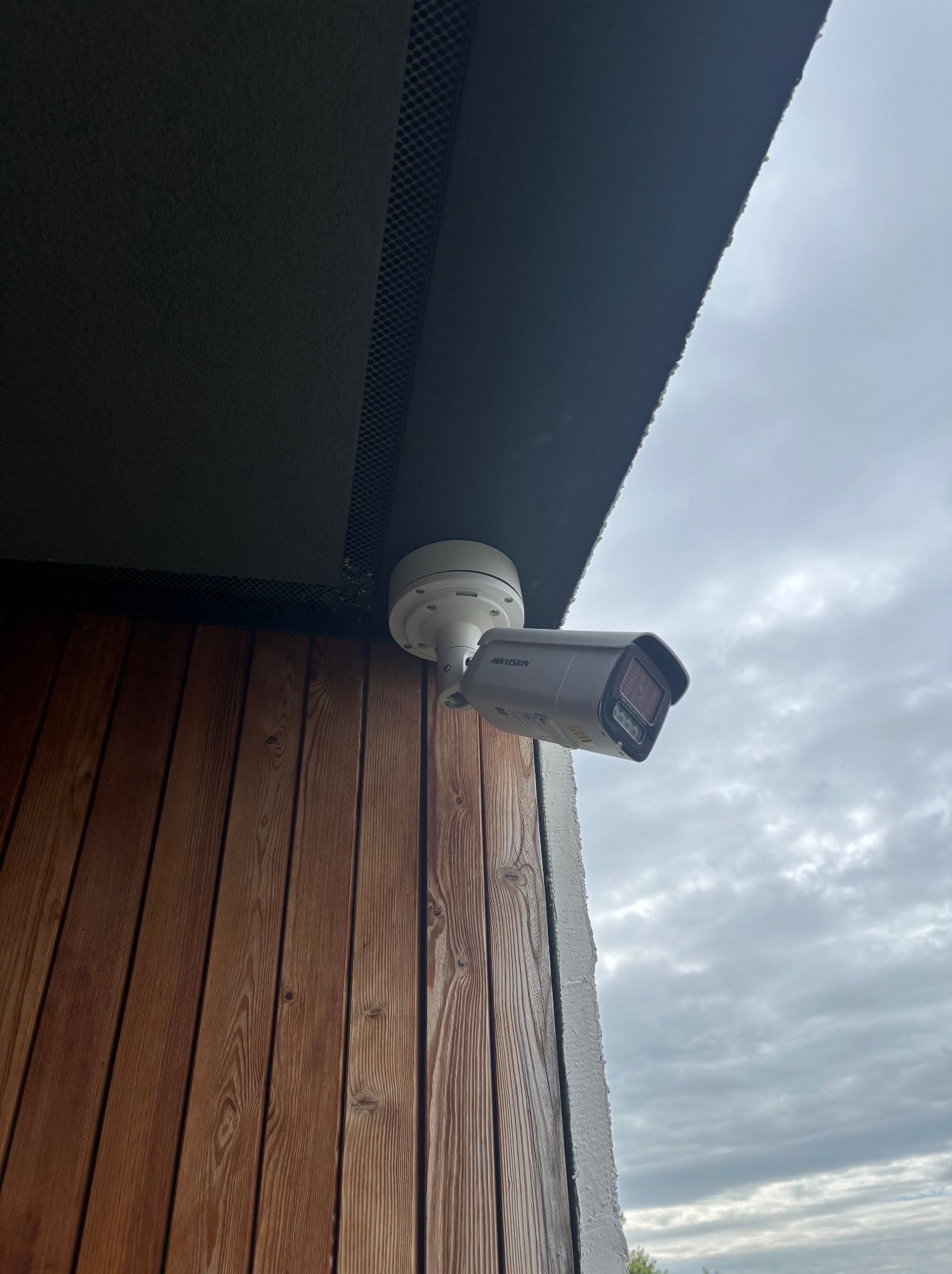Kamera IP Hikvision, zamontowana na drewnianej elewacji budynku w Jasieniu. Widok od dołu, pochmurne niebo w tle. Ochrona domu.