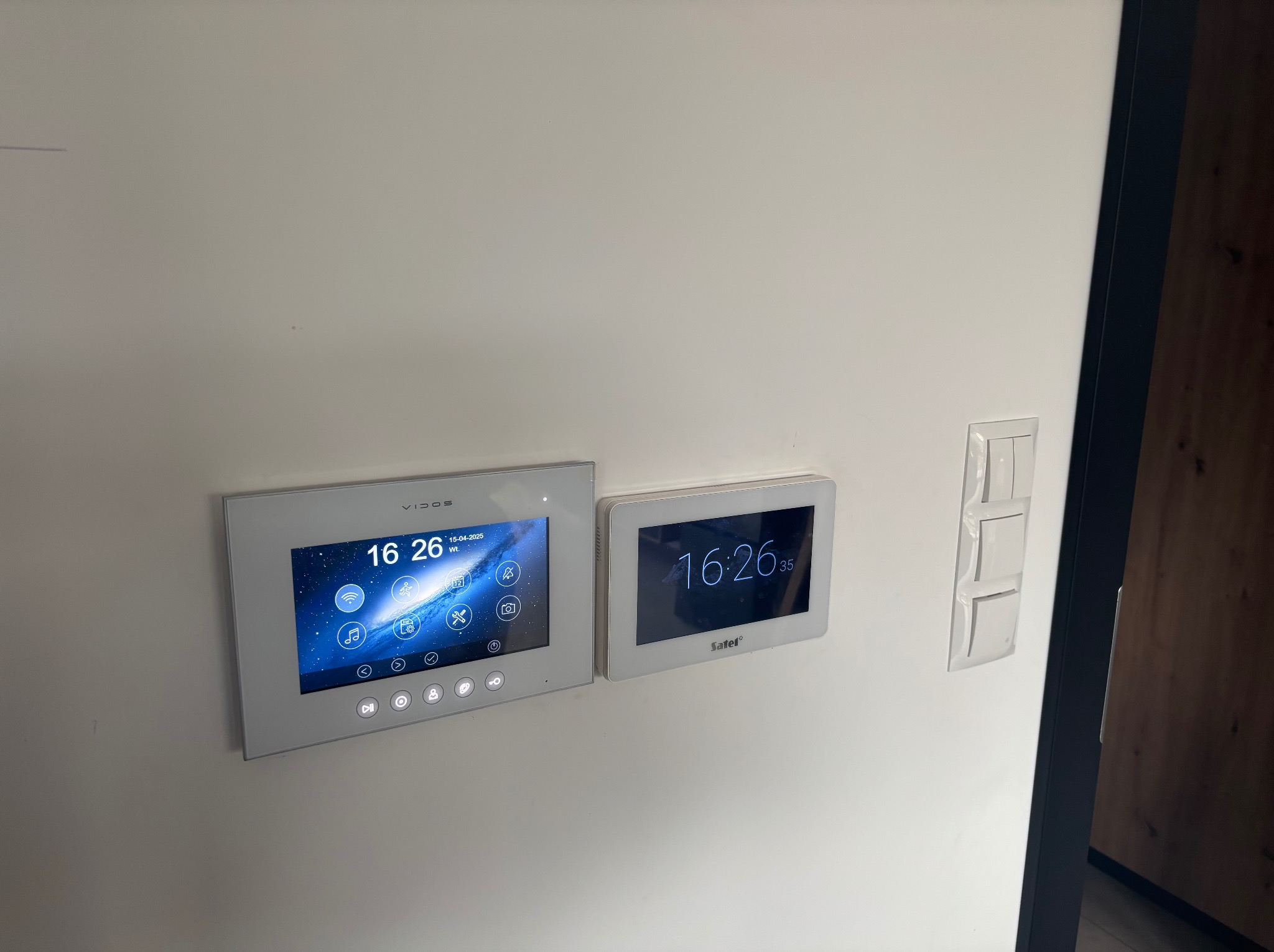 Dwa panele sterowania domofonem Vidos i Satei zamontowane na białej ścianie, obok włącznik światła i fragment czarnej futryny drzwiowej. Ekrany pokazują godzinę 16:26.