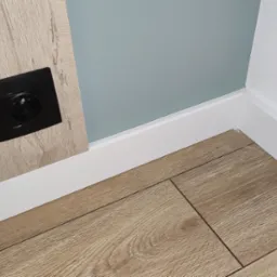 Fragment wnętrza: panele podłogowe w odcieniach brązu, biała listwa przypodłogowa, ściana w kolorze jasnego błękitu i biała, oraz czarne gniazdko elektryczne na drewnopodobnej płycie.