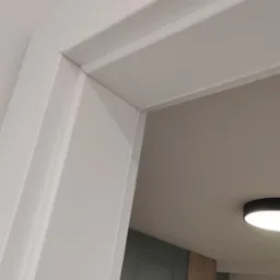 Fragment białej ościeżnicy drzwiowej z widocznym połączeniem z sufitem, w tle zielone drzwi i fragment drewnianej okładziny ściennej.