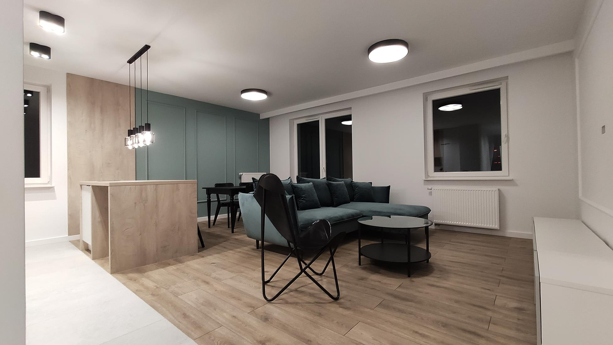 Jasne wnętrze salonu z drewnianą podłogą, turkusową sofą narożną, czarnym stolikiem kawowym i krzesłem typu motyl. Widoczne panele ścienne w kolorze zielonym i beżowym. Nad blatem kuchennym wiszą...