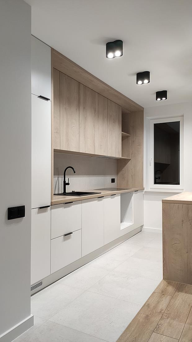 Nowoczesna kuchnia z białymi szafkami dolnymi i drewnopodobnymi szafkami górnymi, czarną armaturą i trzema czarnymi lampami sufitowymi