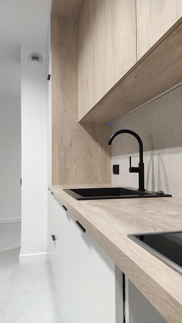 Nowoczesna kuchnia z czarną armaturą i blatem imitującym jasne drewno, białe fronty szafek z minimalistycznymi uchwytami, widok na zlew i fragment płyty indukcyjnej.