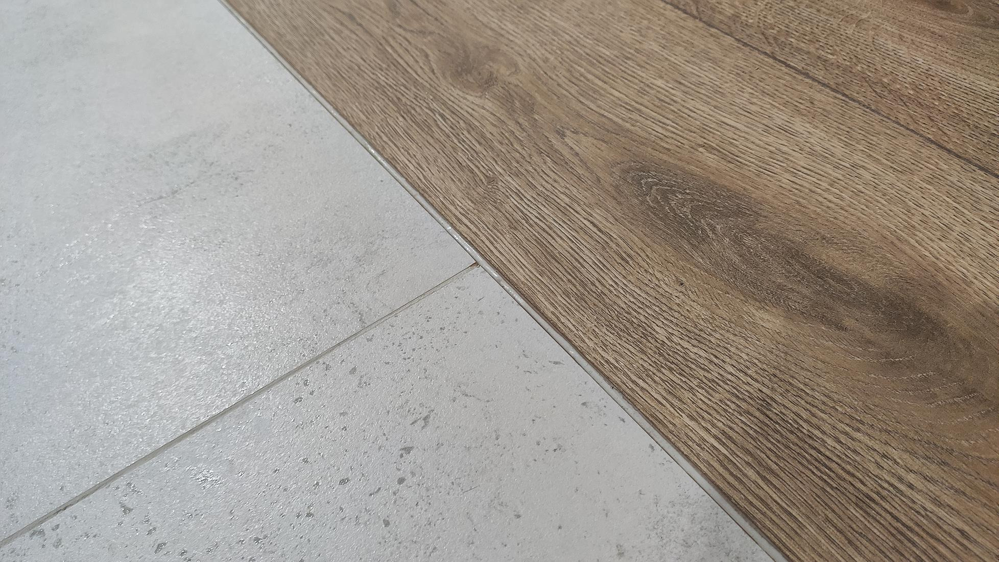 Połączenie jasnych, matowych płytek podłogowych imitujących kamień z panelami podłogowymi w odcieniach drewna. Widoczna fuga między płytkami.