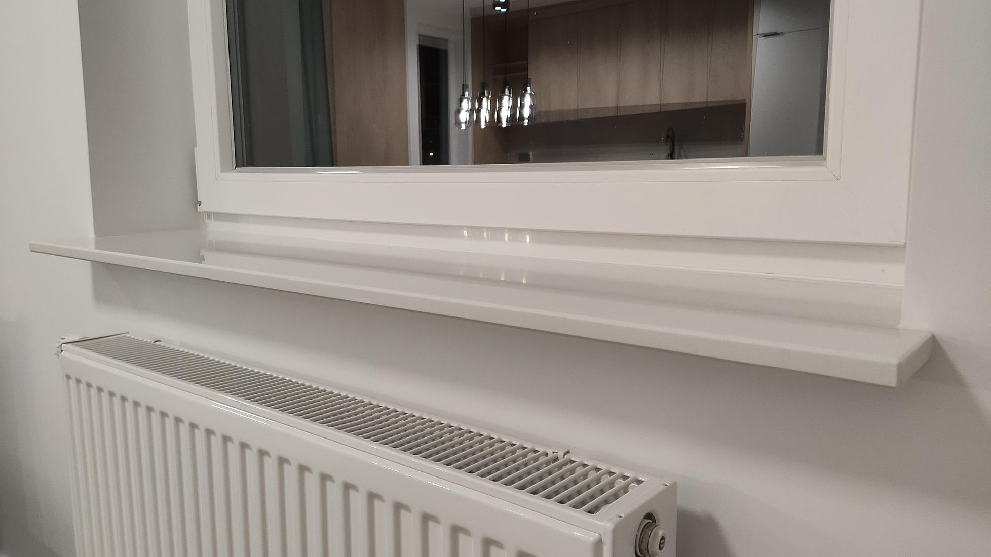Białe okno z parapetem nad grzejnikiem, widok na kuchnię z nowoczesnym oświetleniem.