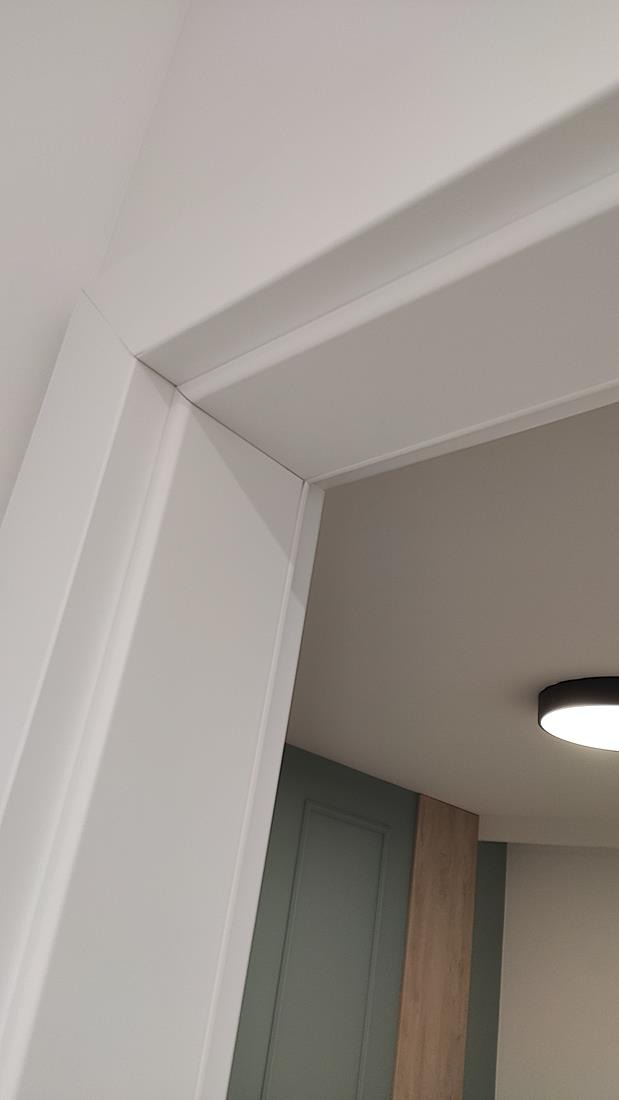 Fragment białej ościeżnicy drzwiowej z widocznym połączeniem z sufitem, w tle zielone drzwi i fragment drewnianej okładziny ściennej.