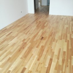 Usługi remontowo budowlane, hydrauliczne i elektryczne. - Jasne, lakierowane deski podłogowe ułożone w jodełkę w pustym, odświeżonym pokoju z białymi ścianami, widok w głąb otwartych drzwi do kolejnych pomieszczeń.