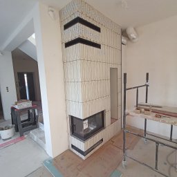 Usługi remontowo budowlane, hydrauliczne i elektryczne. - Wnętrze w Chełmie z kominkiem obłożonym beżowymi płytkami w pionowy wzór, czarne akcenty. Widoczne rusztowanie i materiały budowlane.