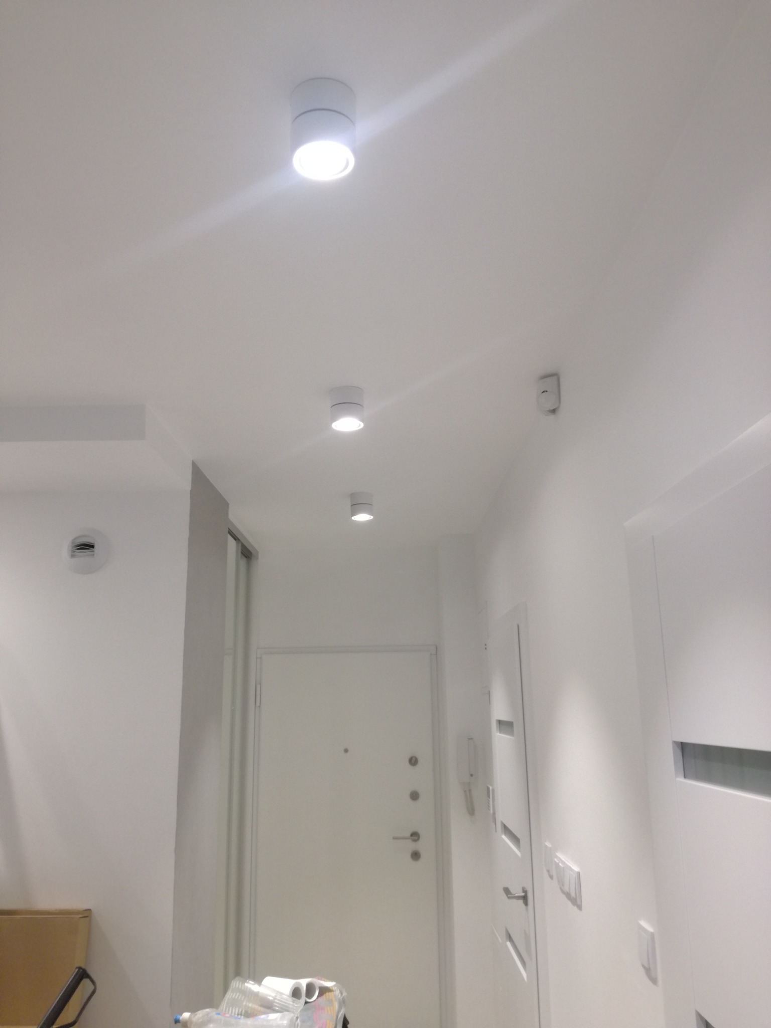 Białe, cylindryczne lampy sufitowe LED w nowocześnie wykończonym, jasnym przedpokoju z białymi ścianami i drzwiami.