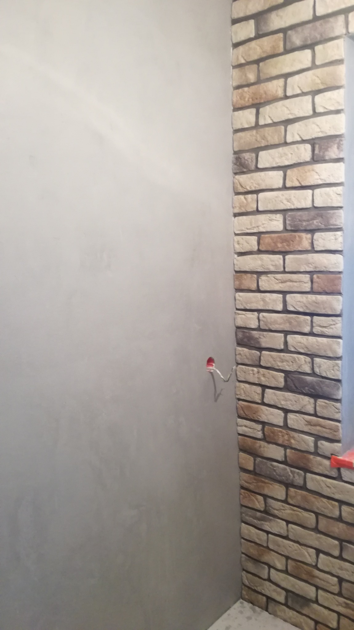 Ściana z surową gładzią kontrastująca z fragmentem ściany wykończonej dekoracyjną okładziną imitującą cegłę, widoczne wystające przewody elektryczne.