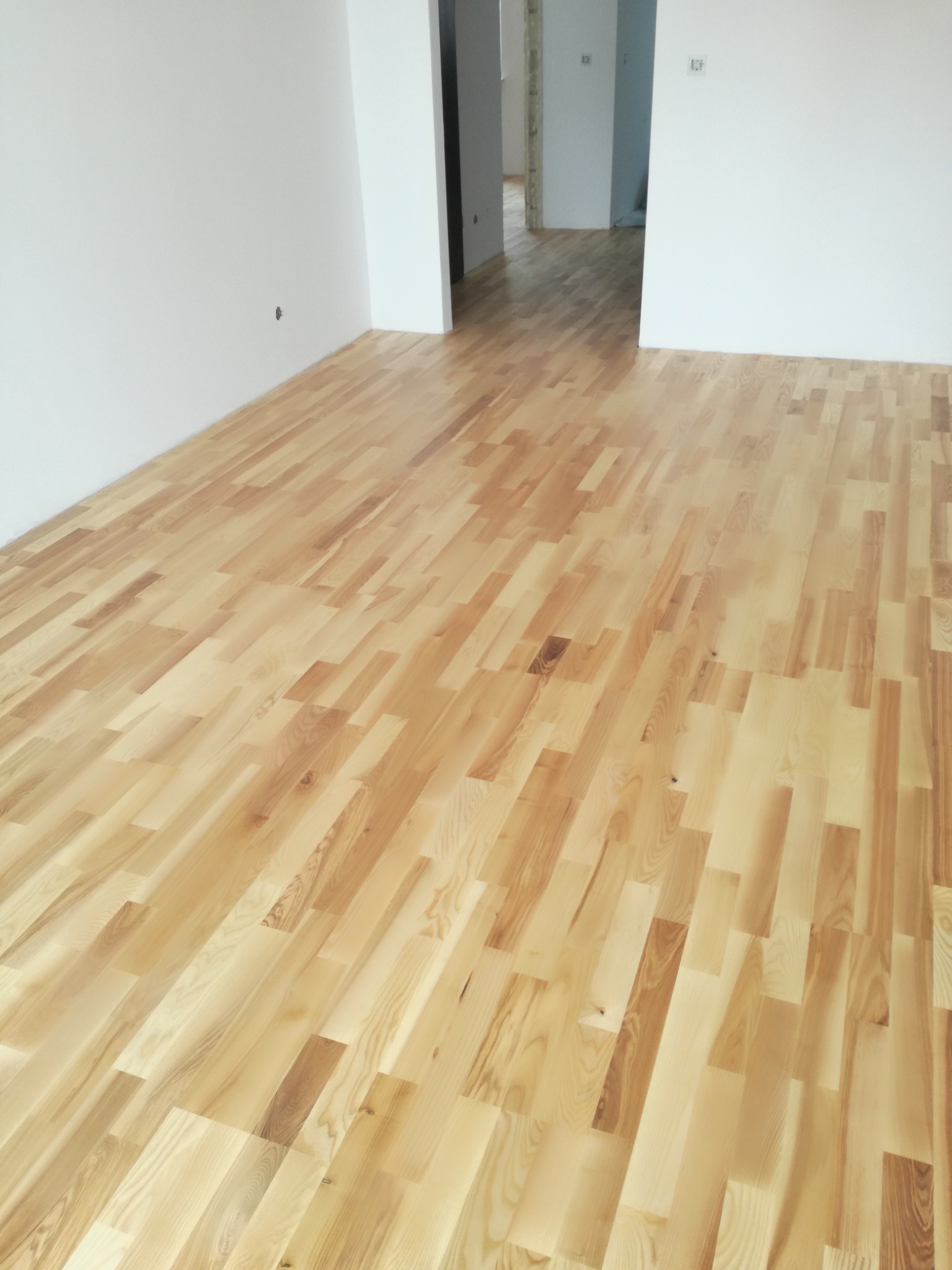 Jasne, lakierowane deski podłogowe ułożone w jodełkę w pustym, odświeżonym pokoju z białymi ścianami, widok w głąb otwartych drzwi do kolejnych pomieszczeń.