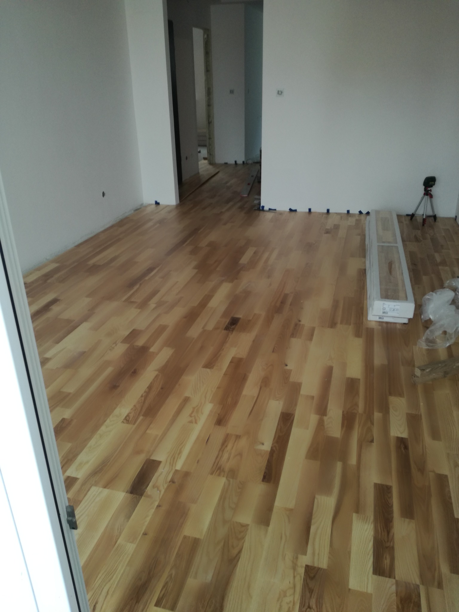 Parkiet z jasnego drewna ułożony w nowym pomieszczeniu, zabezpieczony taśmą malarską przy ścianach, z laserowym niwelatorem na trójnogu i paczkami paneli w rogu.