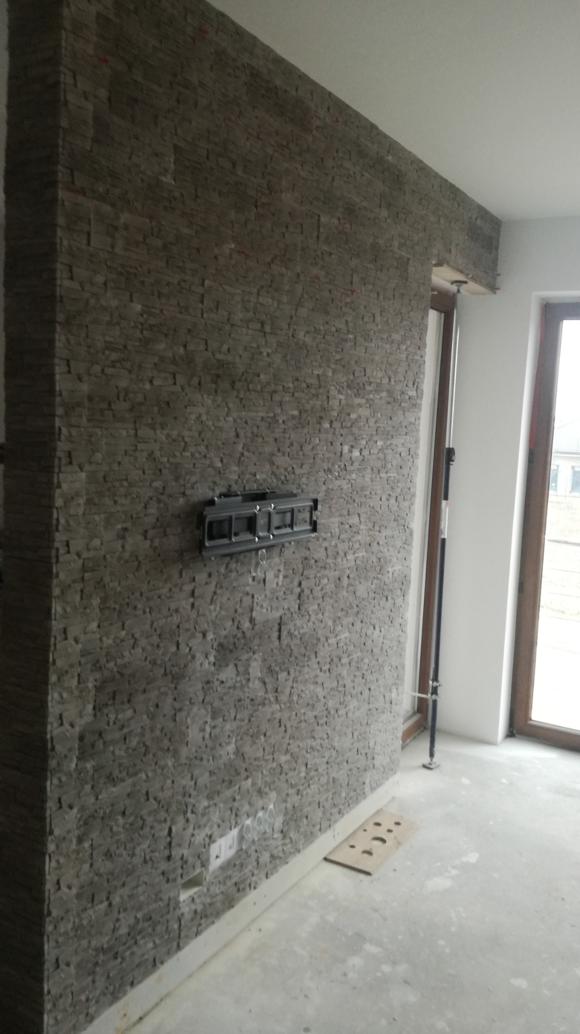 Ściana wykończona kamieniem dekoracyjnym w odcieniach szarości, z zamontowanym uchwytem do telewizora i widocznymi gniazdkami elektrycznymi. Fragment okna z drewnianą ramą po prawej stronie.