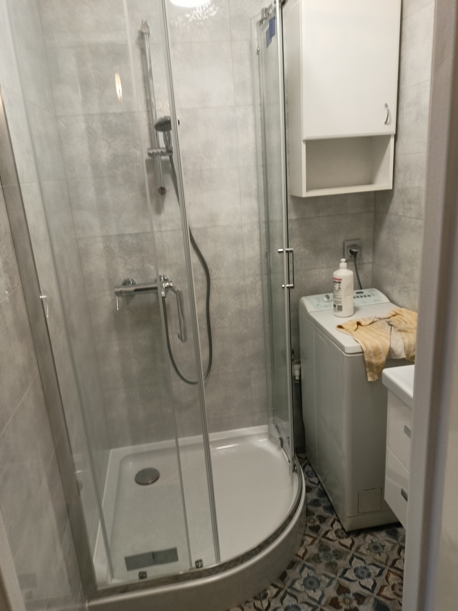 Wnętrze łazienki po remoncie: kabina prysznicowa narożna z szarymi płytkami imitującymi kamień, biała szafka wisząca nad pralką z ręcznikiem, oraz wzorzysta podłoga.