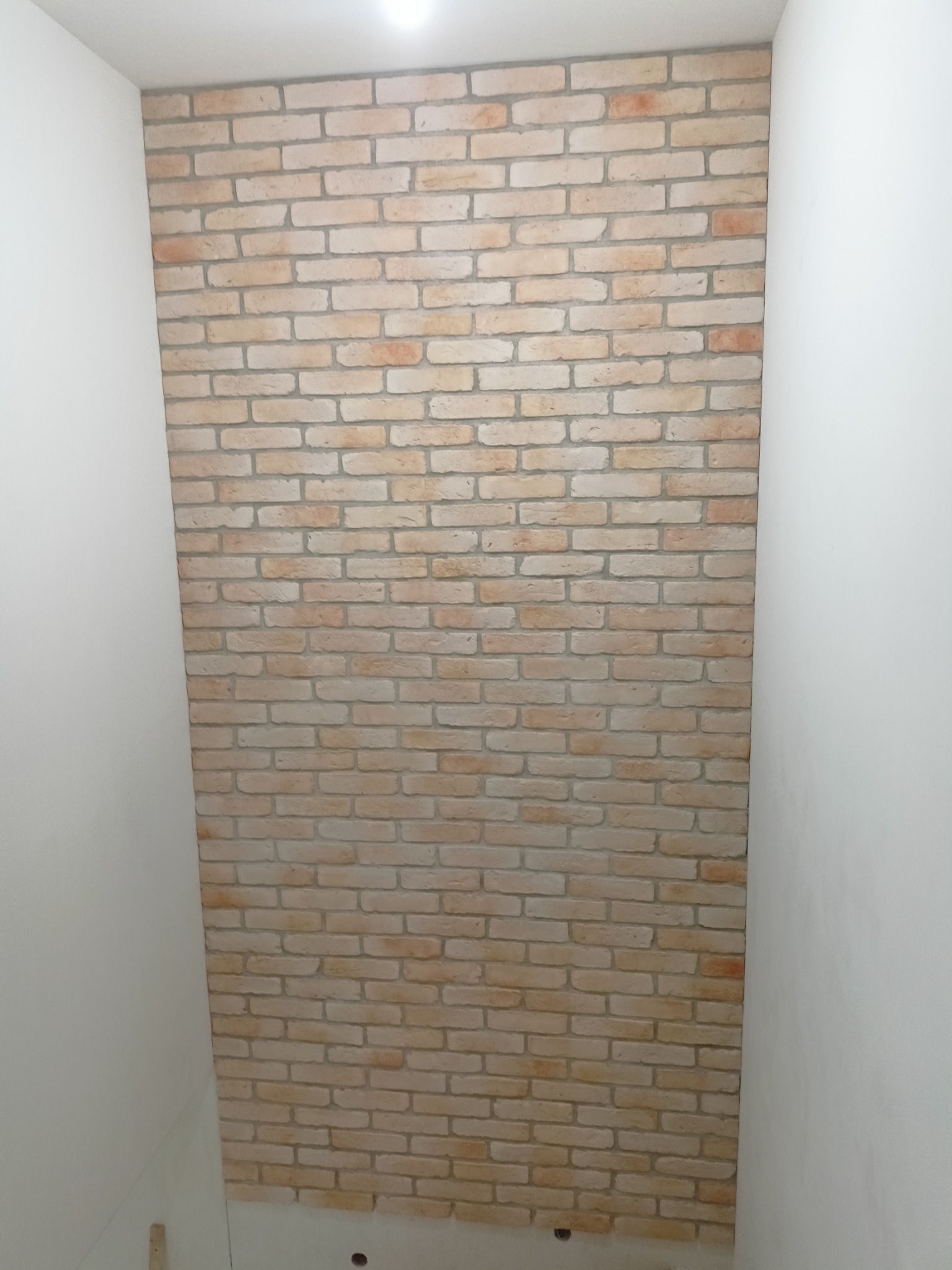Ściana wykończona dekoracyjną cegłą w odcieniach beżu i brązu, obok gładka, biała ściana, widoczne niedoskonałości fug i drobne ubytki w niektórych cegłach, tworzące rustykalny efekt.