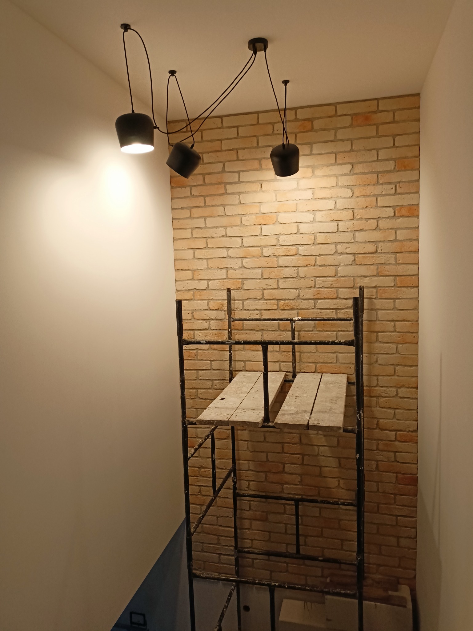 Ściana z cegieł dekoracyjnych, oświetlona trzema nowoczesnymi lampami wiszącymi, z widocznym rusztowaniem malarskim.