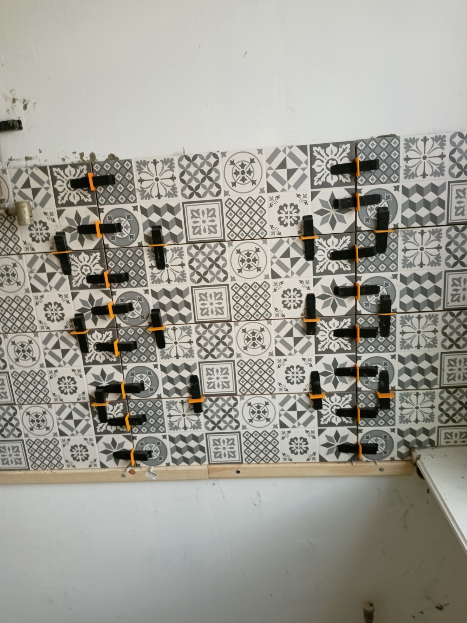 Świeżo ułożone, wzorzyste płytki ścienne w odcieniach szarości, utrzymywane w równych odstępach za pomocą pomarańczowych i czarnych klipsów dystansowych, z widoczną drewnianą listwą podtrzymującą...