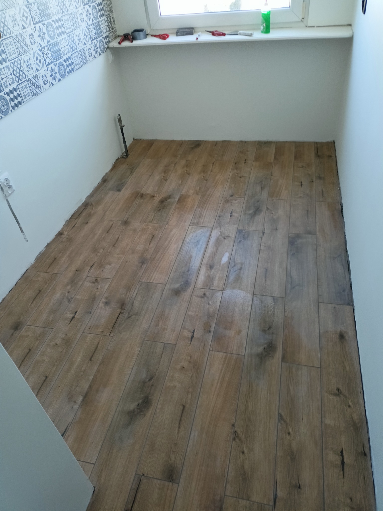 Ułożona podłoga z płytek imitujących drewno w pomieszczeniu z częściowo wykończonymi ścianami i oknem.