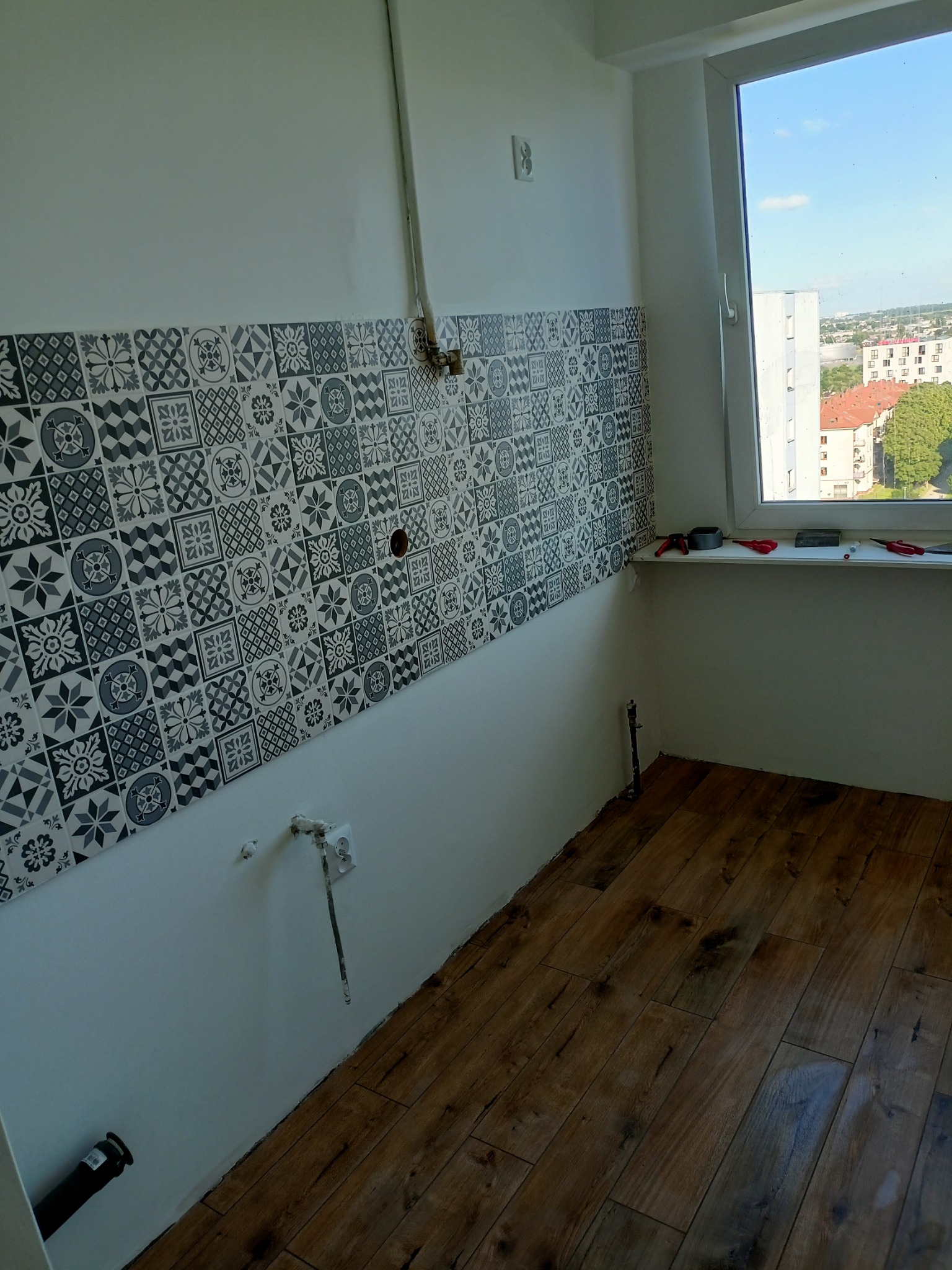 Fragment kuchni w trakcie wykańczania: położone płytki podłogowe imitujące drewno, ściana z częściowo położonymi dekoracyjnymi płytkami ściennymi w odcieniach szarości, wystające rury i gniazdka...