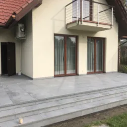 Nowoczesny taras z szarych płyt betonowych, prowadzący do domu z balkonem i brązowymi oknami, widoczne kwiaty w donicach po lewej stronie.