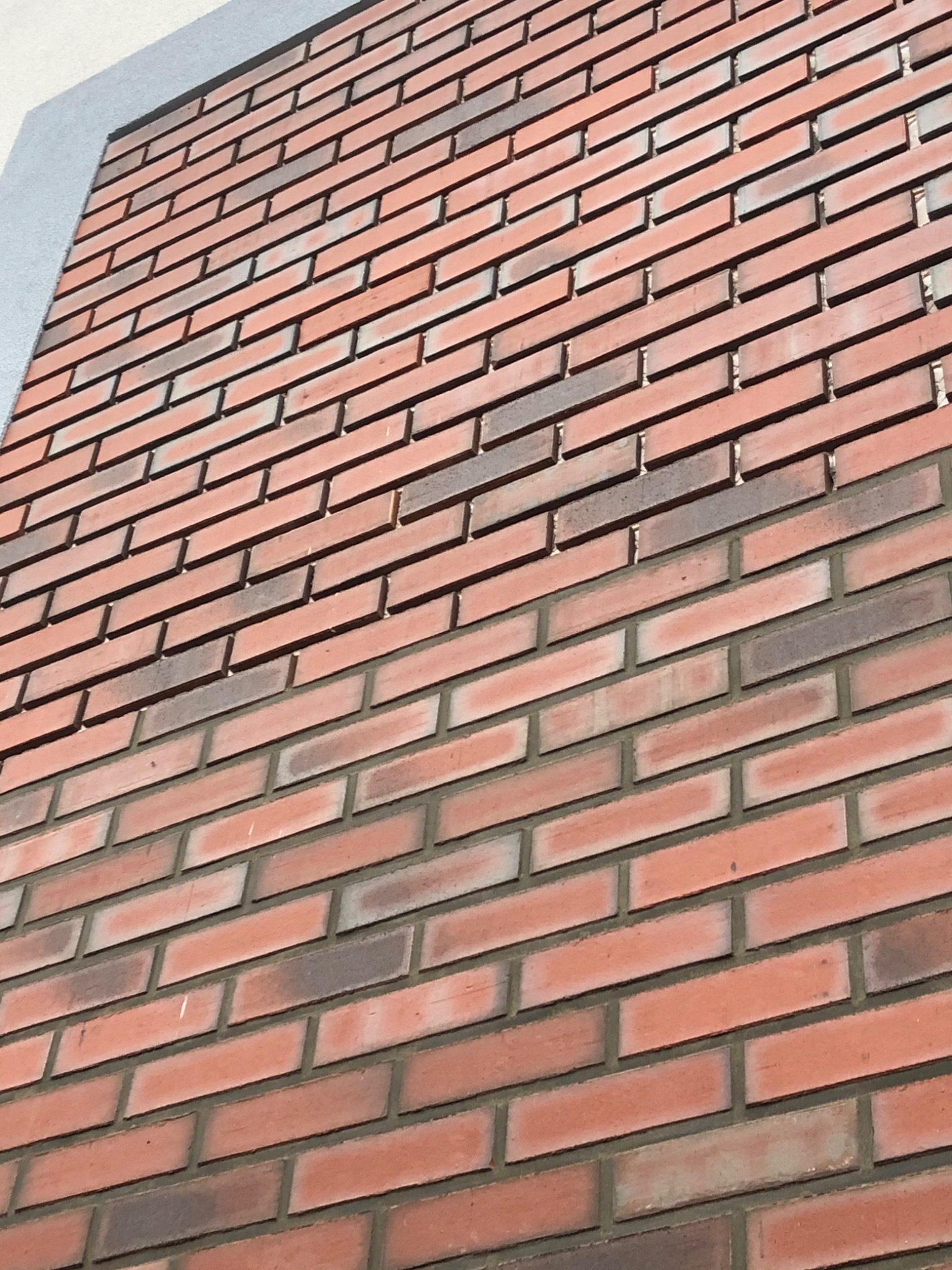 Czerwona ściana z cegły klinkierowej z szarą obwódką, ułożona pod kątem, z widocznymi spoinami i drobnymi przebarwieniami na niektórych cegłach.