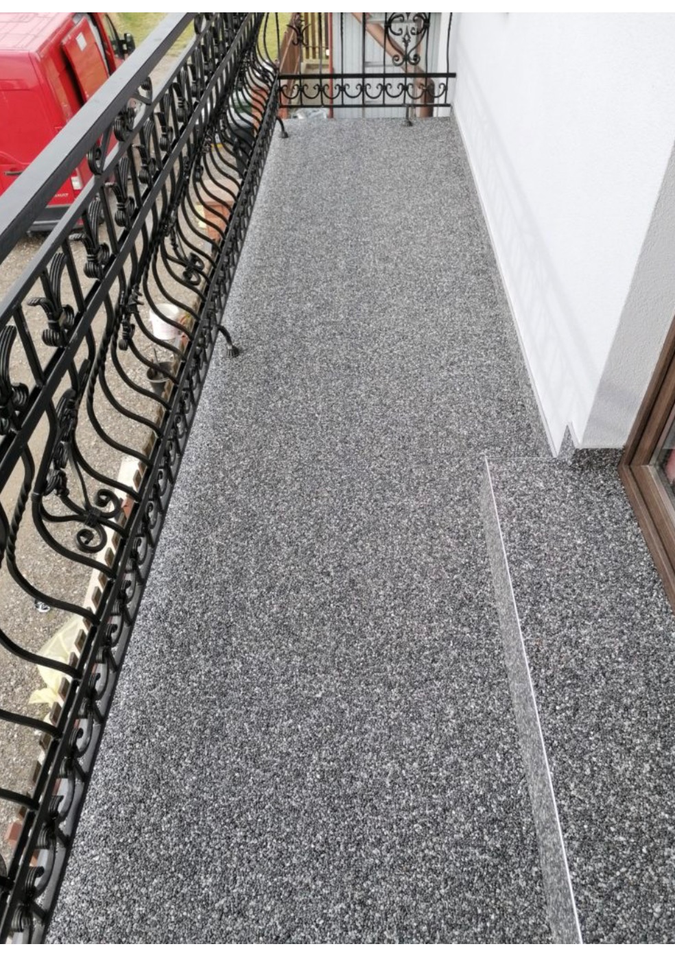 Balkon z szarą, kamyczkową posadzką kompozytową, ozdobną, metalową balustradą i fragmentem białej ściany budynku.