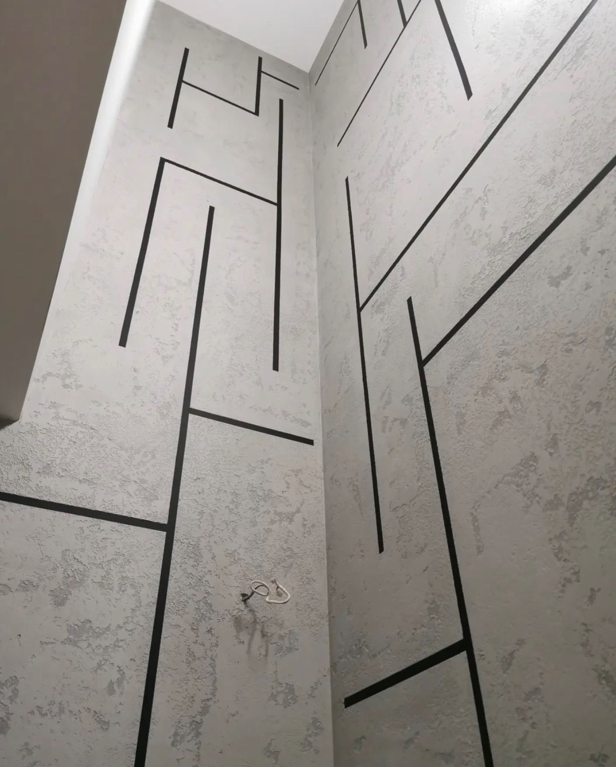 Dekoracyjna ściana we Wrocławiu z szarą strukturą i czarnymi, geometrycznymi liniami, tworzącymi nowoczesny wzór. Na ścianie widoczny wystający kabel.