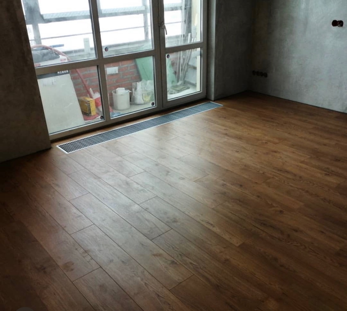 Ułożone panele podłogowe w odcieniach brązu w pokoju z dużym oknem balkonowym i surowymi ścianami, widoczna kratka wentylacyjna przy oknie.