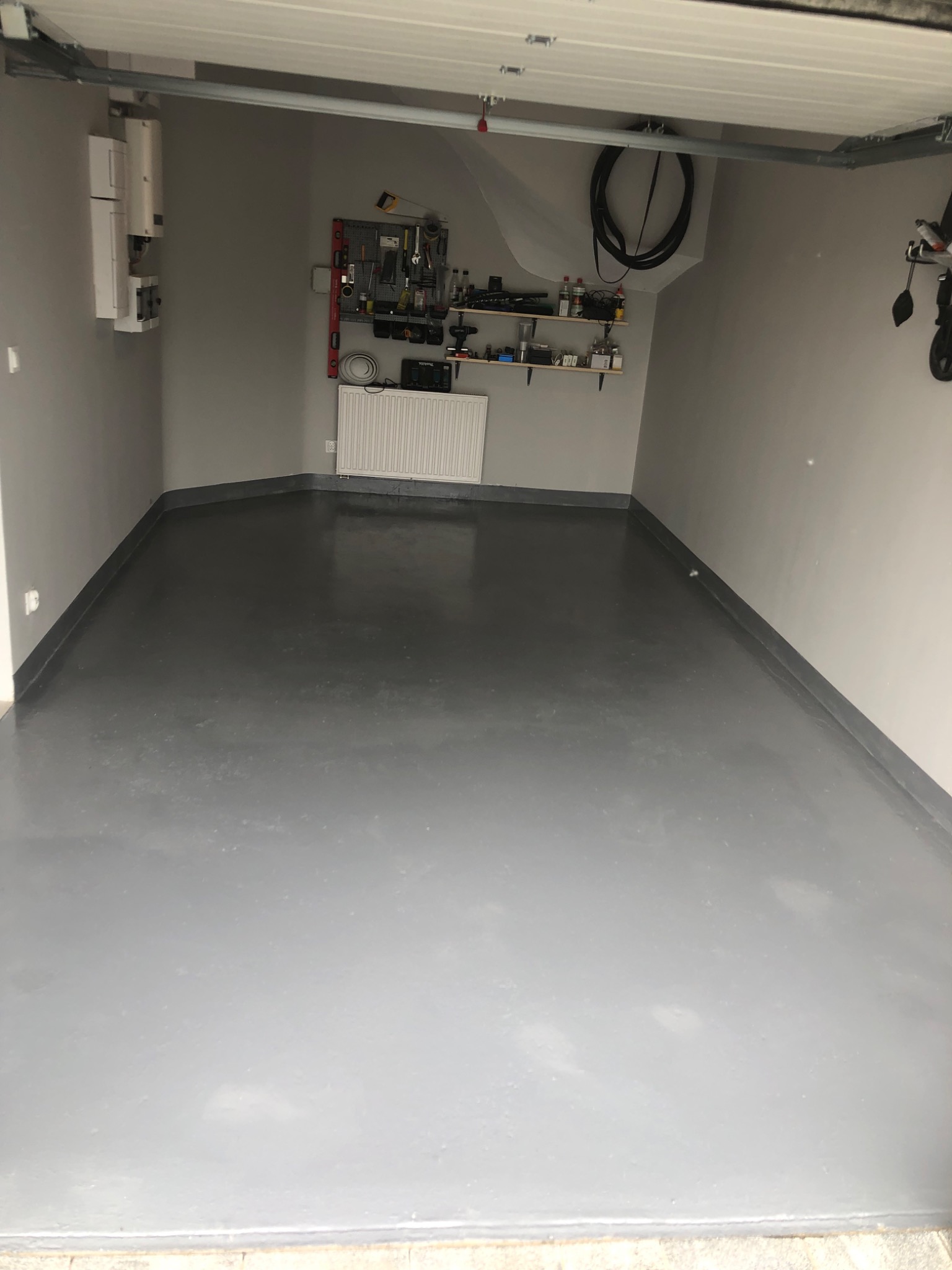 Wnętrze garażu z szarą, gładką posadzką żywiczną, ściany pomalowane na szaro, widoczny grzejnik, panel narzędziowy i instalacja elektryczna.