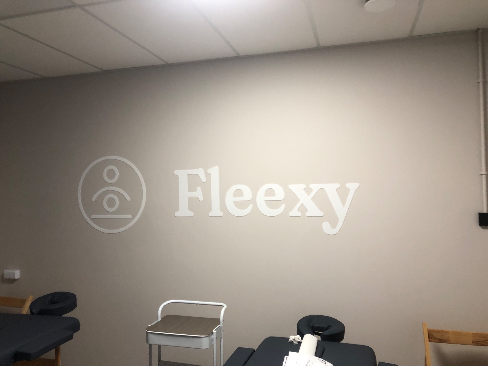 Ściana w gabinecie z logo 'Fleexy' nad profesjonalnym łóżkiem do masażu i wózkiem na akcesoria.
