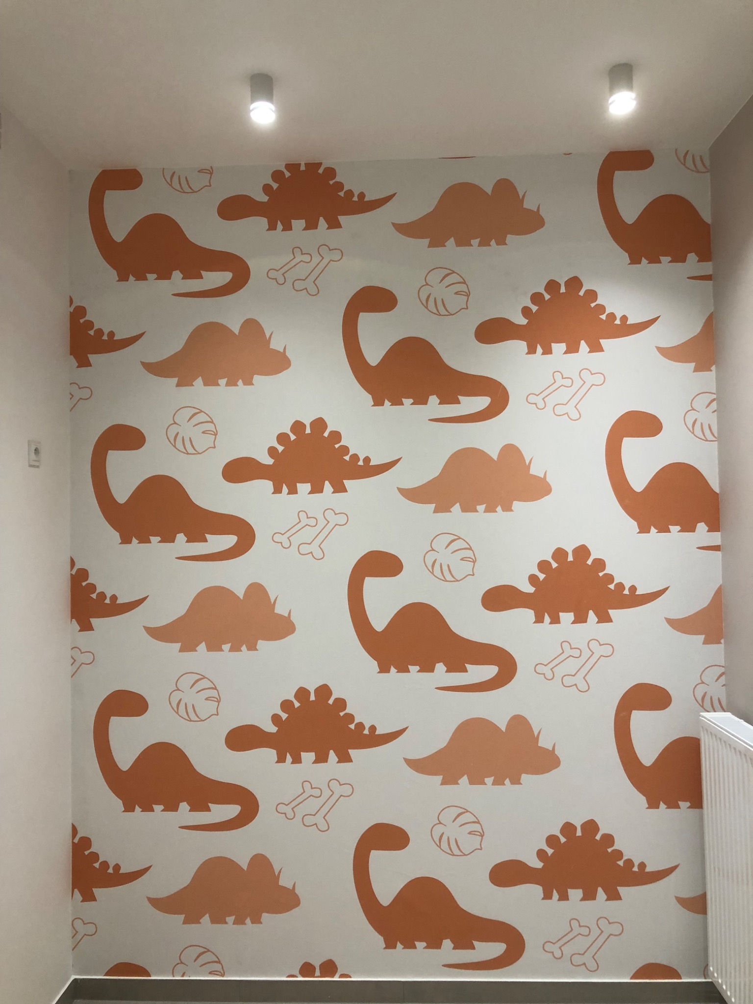 Ściana w pokoju dziecięcym pokryta wzorem w pomarańczowe dinozaury i kości na białym tle, oświetlona przez dwa reflektory sufitowe.